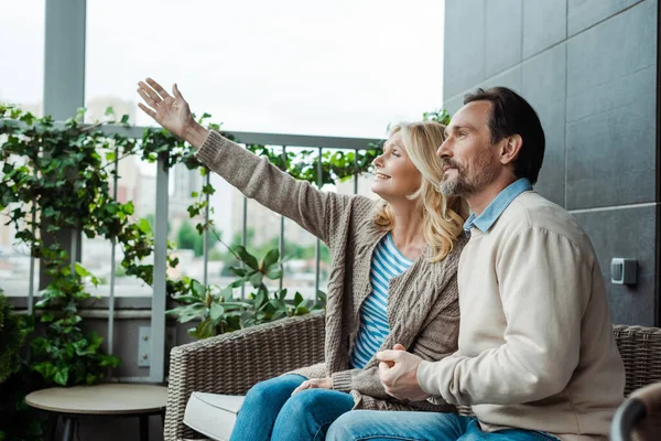 Lächelnde Frau zeigt mit der Hand in die Nähe ihres hübschen Mannes auf dem Weidensofa auf der Terrasse — Stockfoto