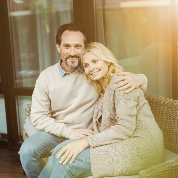 Imagen tonificada de un hombre maduro sonriente abrazando a una hermosa esposa en un sofá de mimbre en la terraza - foto de stock