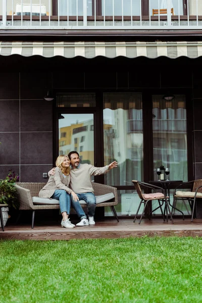 Mature man pointing with hand and embracing smiling wife on wicker sofa on terrace — Stock Photo