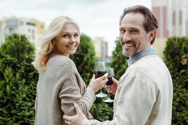 Vue latérale du couple mature souriant à la caméra tout en tenant des verres de vin dans la rue urbaine — Photo de stock