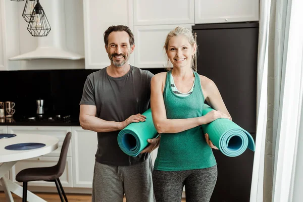 Casal maduro em sportswear sorrindo para a câmera enquanto segurando tapetes de fitness em casa — Fotografia de Stock