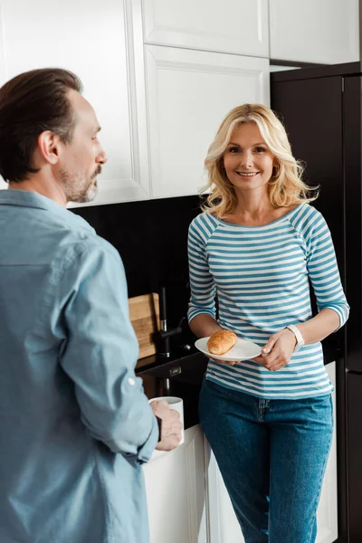 Concentration sélective de la femme souriante tenant croissant près du mari avec tasse de café dans la cuisine — Photo de stock