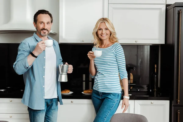 Улыбающаяся взрослая пара смотрит в камеру и пьет кофе на кухне. — стоковое фото