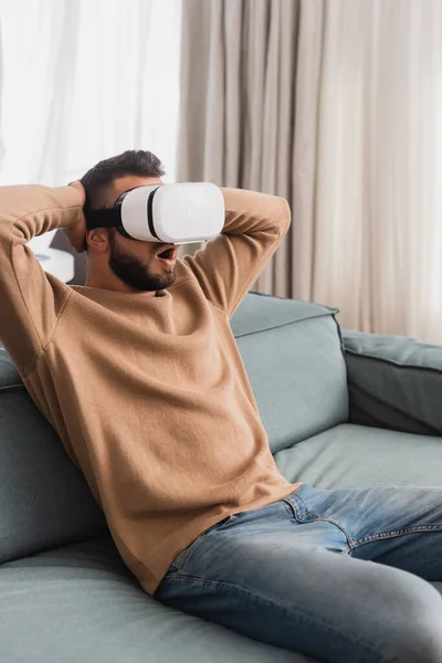 Hombre barbudo sorprendido en auriculares de realidad virtual en la sala de estar - foto de stock