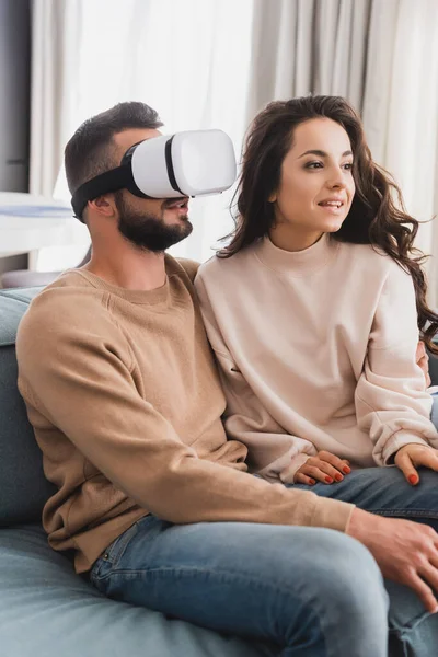 Hermosa chica sentada en el sofá cerca de novio barbudo en auriculares de realidad virtual - foto de stock