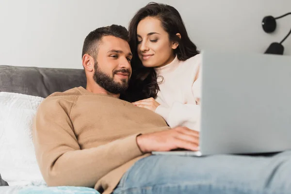 Enfoque selectivo de freelancer feliz utilizando el ordenador portátil cerca de hermosa novia en el dormitorio - foto de stock