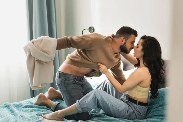 Side view of passionate man holding sweatshirt of attractive girlfriend in bedroom — Stock Photo