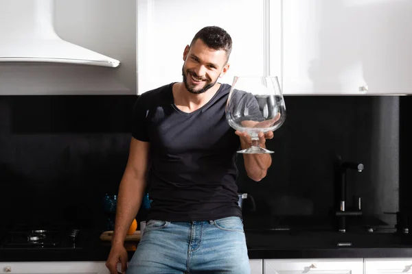 Веселий бородатий чоловік тримає величезний келих вина на кухні — стокове фото