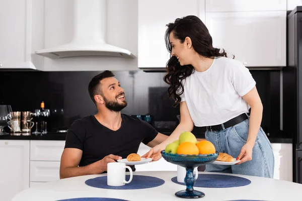 Mujer alegre sosteniendo platos con cruasanes cerca novio feliz en la cocina - foto de stock