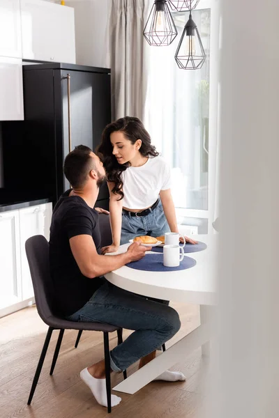 Селективное внимание привлекательной женщины, смотрящей на красивого парня рядом с завтраком на кухне — стоковое фото