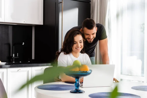 Selective focus of happy man looking at laptop near girlfriend and tasty fruits — Stock Photo