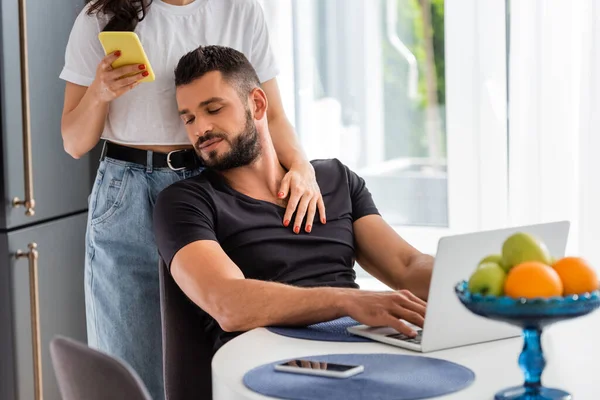 Selective focus of woman standing and touching bearded freelancer boyfriend near laptop while using smartphone — Stock Photo