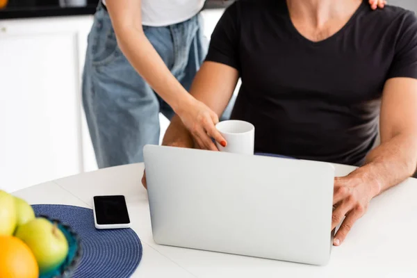 Обрезанный вид женщины касаясь чашки рядом с фрилансером бойфренд, ноутбук и смартфон с чистым экраном — стоковое фото
