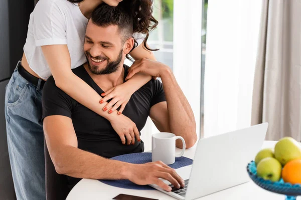 Selektiver Fokus der jungen Frau, die ihren glücklichen Freund in der Nähe des Laptops umarmt — Stockfoto