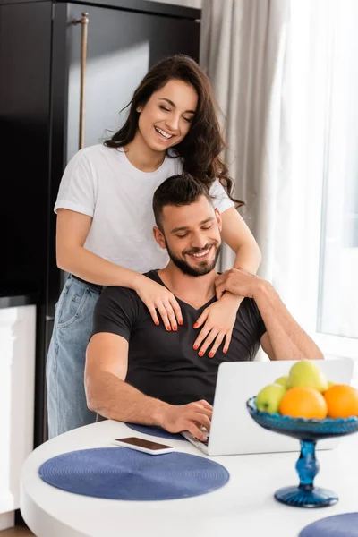 Selective focus of happy couple looking at laptop near smartphone with blank screen — Stock Photo