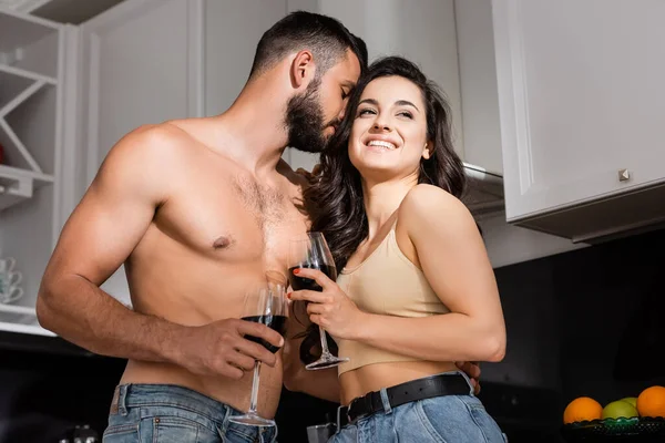 Vista de ángulo bajo del hombre sin camisa oliendo el pelo de la joven feliz sosteniendo un vaso de vino tinto - foto de stock