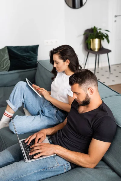 Barbudo freelancer utilizando portátil cerca de novia con tableta digital en la sala de estar - foto de stock