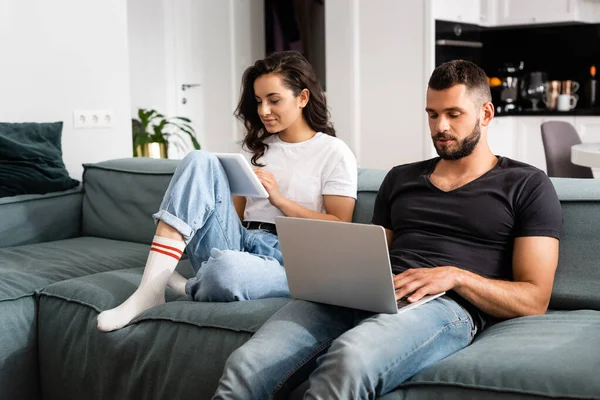 Freelancer bonito usando laptop perto da namorada com tablet digital na sala de estar — Fotografia de Stock