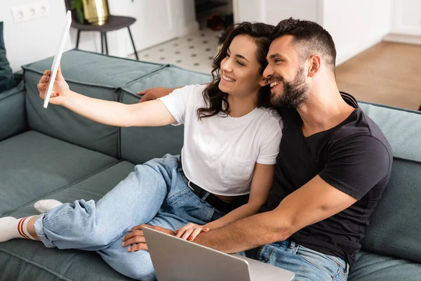Chica feliz sosteniendo la tableta digital y tomando selfie con novio guapo cerca de la computadora portátil - foto de stock