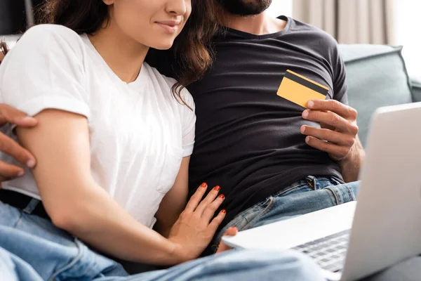 Visão cortada de homem barbudo segurando cartão de crédito e abraçando namorada feliz perto de laptop — Fotografia de Stock