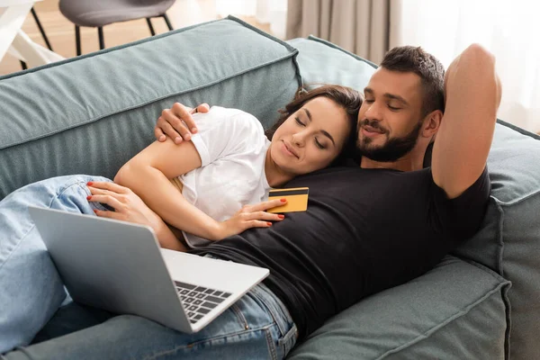 Счастливая женщина лежит на диване с красивым парнем и держит кредитную карту возле ноутбука — стоковое фото