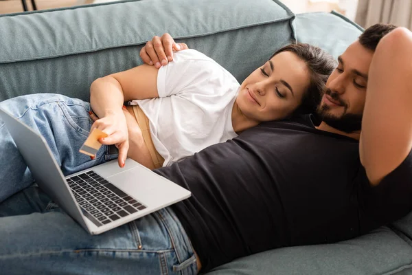 Jolie femme couchée sur le canapé avec beau petit ami et tenant la carte de crédit près d'un ordinateur portable — Photo de stock