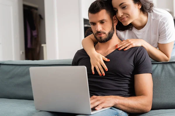 Chica feliz abrazando novio usando el ordenador portátil en la sala de estar - foto de stock