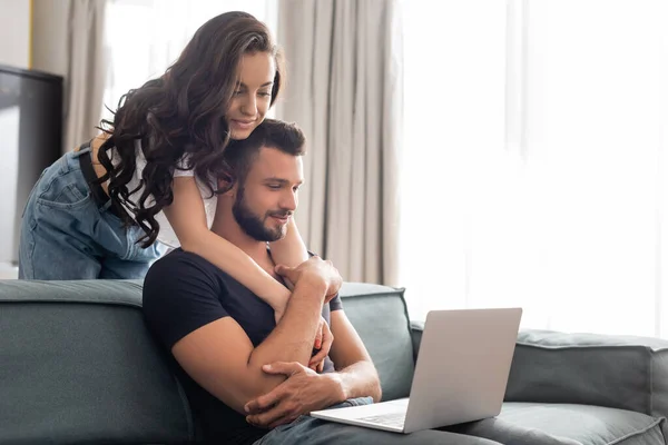Chica feliz abrazando novio guapo usando portátil en la sala de estar - foto de stock