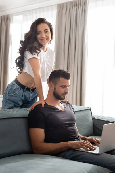 Menina feliz sorrindo perto bonito namorado usando laptop na sala de estar — Fotografia de Stock