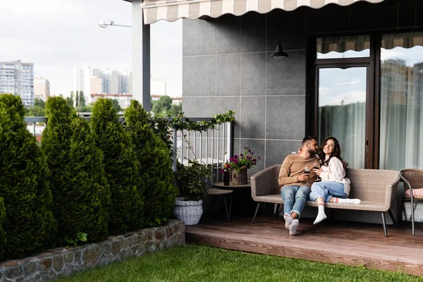 Homem bonito e mulher feliz sentado no sofá ao ar livre e segurando copos de vinho — Fotografia de Stock