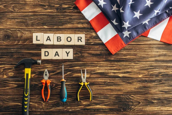 Vista superior de la bandera americana cerca de instrumentos y cubos con letras del día del trabajo en la superficie de madera - foto de stock