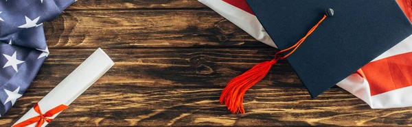 Cultivo horizontal de la gorra de graduación y diploma cerca de bandera americana con estrellas y rayas en la superficie de madera - foto de stock