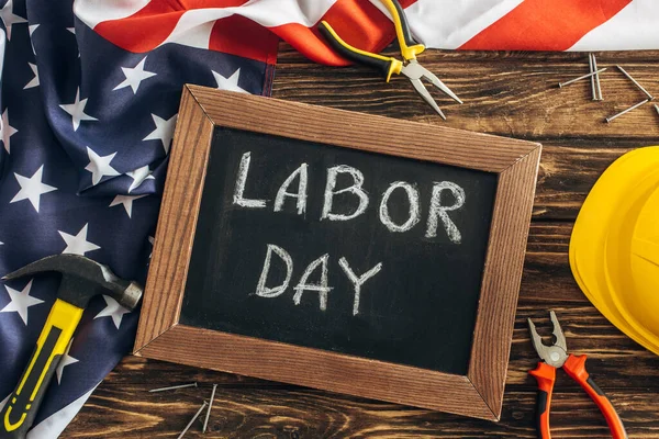 Vista superior de la bandera americana y los instrumentos cerca de pizarra con letras del día del trabajo en la superficie de madera - foto de stock