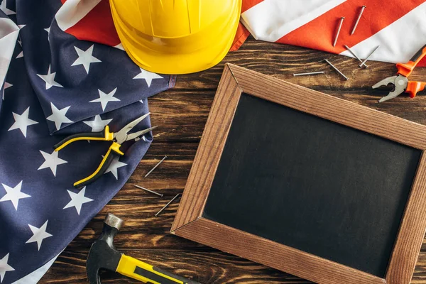 Вид сверху на американский флаг и инструменты рядом с пустой доской на деревянной поверхности, концепция трудового дня — стоковое фото