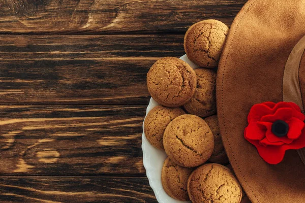 Vista superior de la flor artificial, sombrero de fieltro y galletas en la superficie de madera, concepto de día anzac - foto de stock