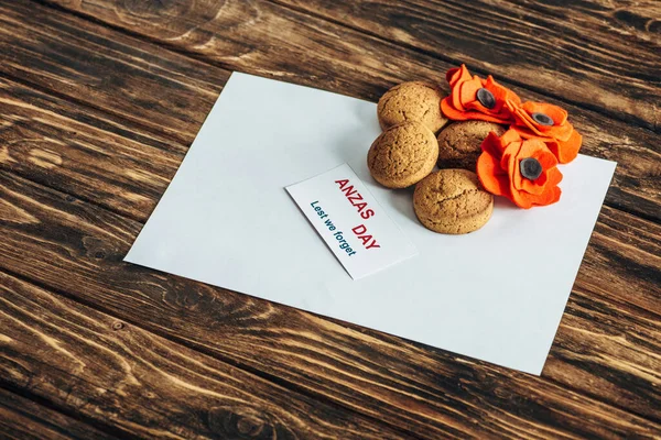 Карточка с анзасом день буквы рядом искусственные цветы, печенье и чистая бумага на деревянной поверхности — стоковое фото