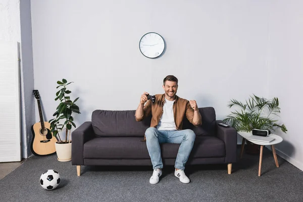 KYIV, UCRANIA - 9 de junio de 2020: hombre feliz mostrando el gesto del ganador mientras está sentado en el sofá y sosteniendo el joystick - foto de stock