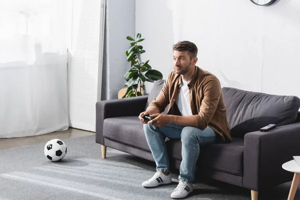 KYIV, UCRAINA - 9 GIUGNO 2020: uomo attento che gioca ai videogiochi seduto sul divano vicino al pallone da calcio — Foto stock