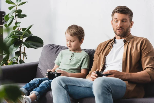 KYIV, UCRANIA - 9 de junio de 2020: padre e hijo atentos sentados en el sofá y jugando videojuegos - foto de stock