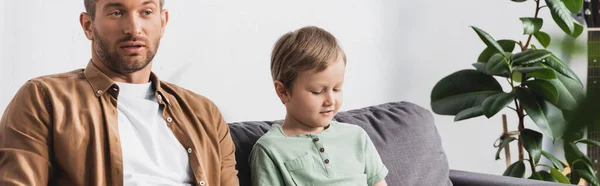 Panoramic crop of thoughtful father and son sitting on sofa near potted plant — Stock Photo