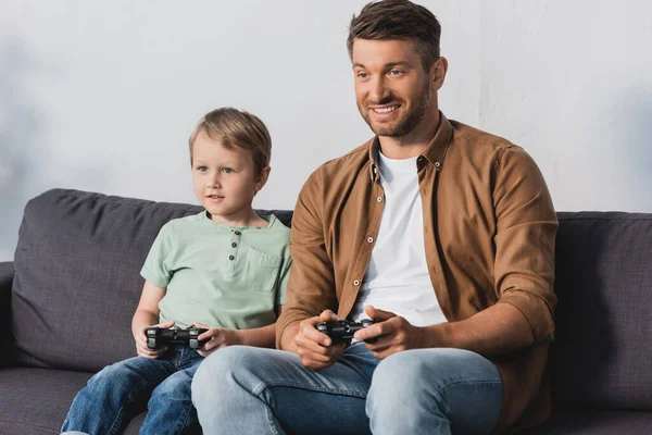 KIEW, UKRAINE - 9. JUNI 2020: Lächelnder Vater und Sohn sitzen auf dem Sofa und spielen Videospiel mit Jostcks — Stockfoto