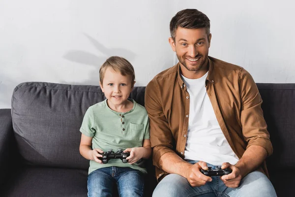KYIV, UCRANIA - 9 de junio de 2020: padre e hijo emocionados mirando a la cámara mientras juegan a videojuegos con joysticks - foto de stock