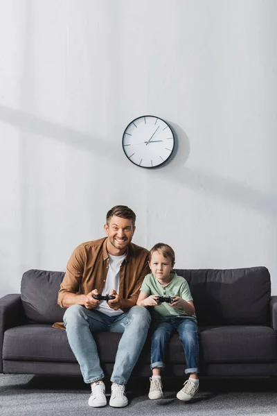 KYIV, UKRAINE - JUNE 9, 2020: happy father and son sitting on sofa and playing video game with joysticks — Stock Photo