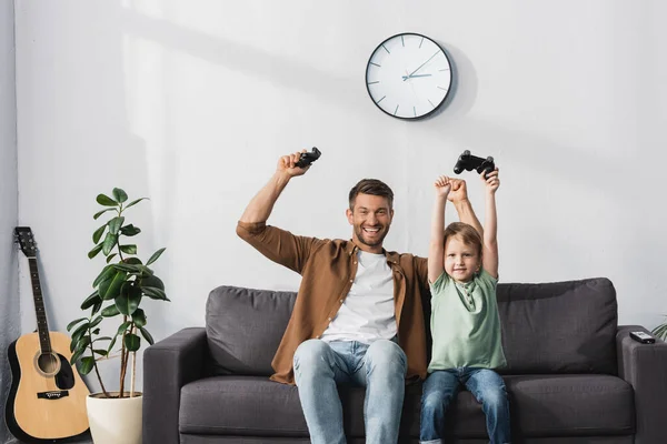 KYIV, UCRANIA - 9 de junio de 2020: feliz padre e hijo sosteniendo joysticks y mostrando gestos ganadores - foto de stock