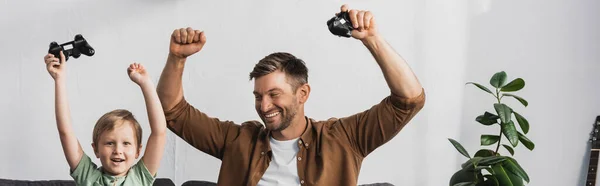 KYIV, UKRAINE - JUNE 9, 2020: horizontal image of excited father and son holding joysticks and showing winner gestures — Stock Photo