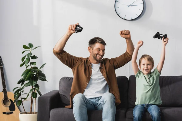 KYIV, UCRANIA - 9 de junio de 2020: feliz padre e hijo mostrando gestos ganadores mientras sostiene joysticks - foto de stock