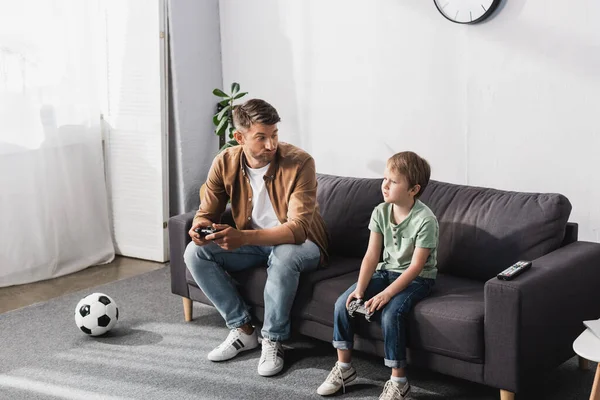 KYIV, UCRANIA - 9 de junio de 2020: padre disgustado mirando a su hijo molesto mientras está sentado en el sofá con joysticks - foto de stock