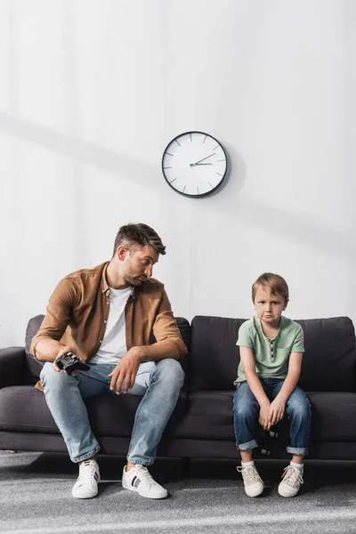 KYIV, UKRAINE - JUNE 9, 2020: upset father looking at sad son while sitting on sofa with joysticks — Stock Photo