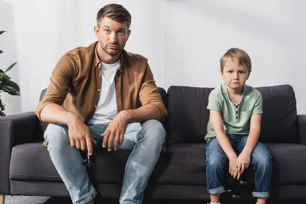 KYIV, UKRAINE - JUNE 9, 2020: upset father and son looking at camera while sitting on sofa with joysticks — Stock Photo