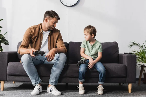 KYIV, UKRAINE - JUNE 9, 2020: upset father and son looking at each other while sitting on sofa with joysticks — Stock Photo